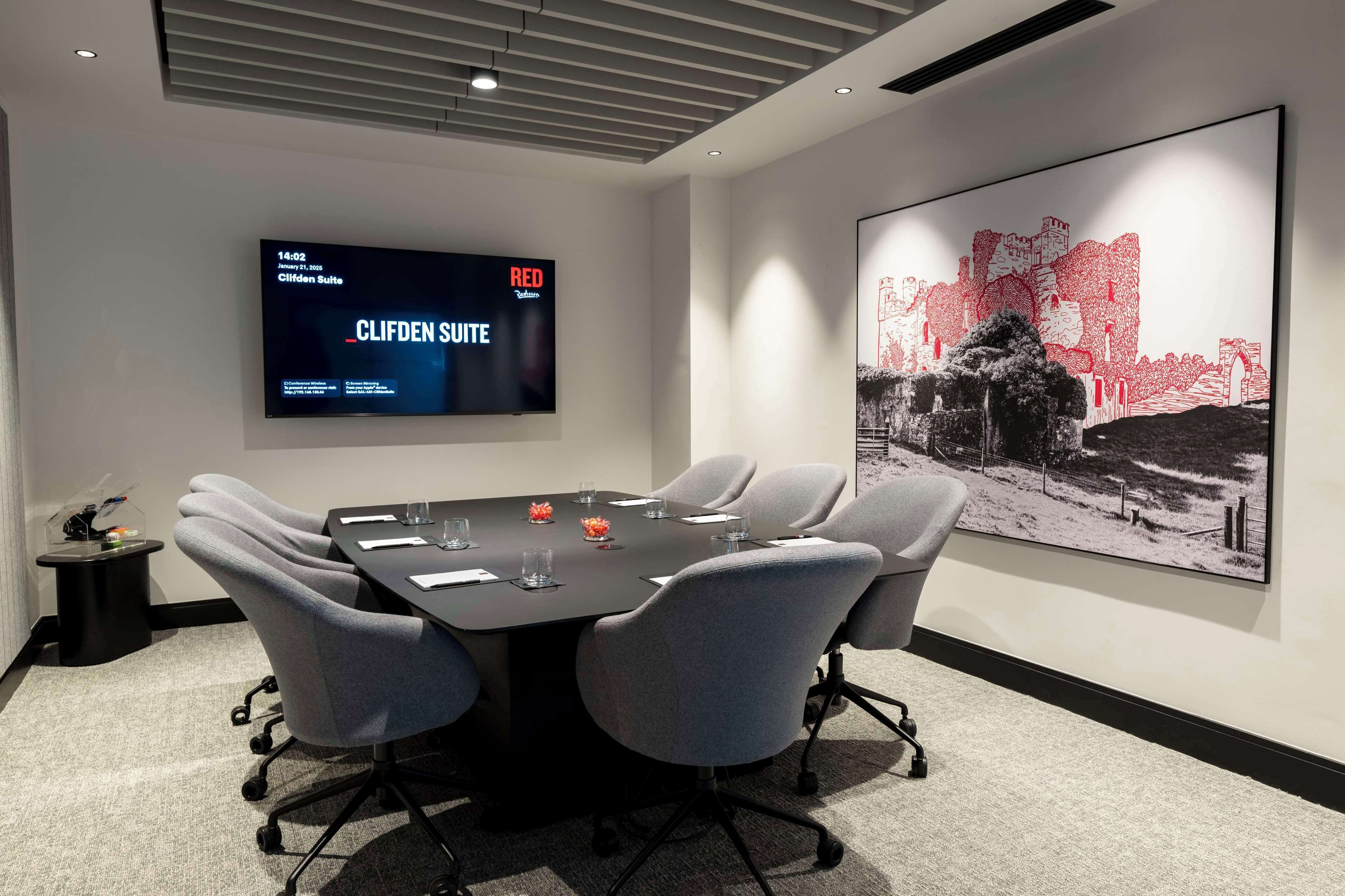 A conference room with a large table, six chairs, a TV screen displaying 'CLIFDEN SUITE', and a large abstract painting on the wall.