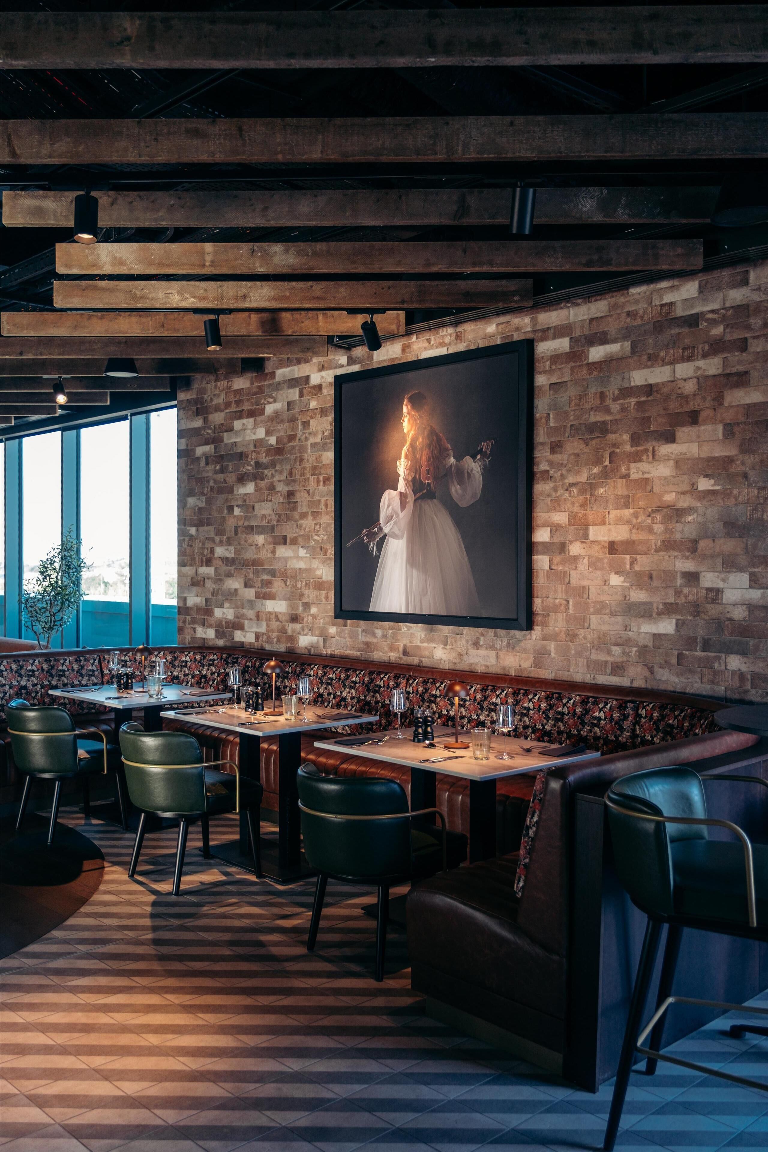A restaurant with a brick wall, wooden ceiling beams, and a large framed portrait. Tables are set with chairs and candles.