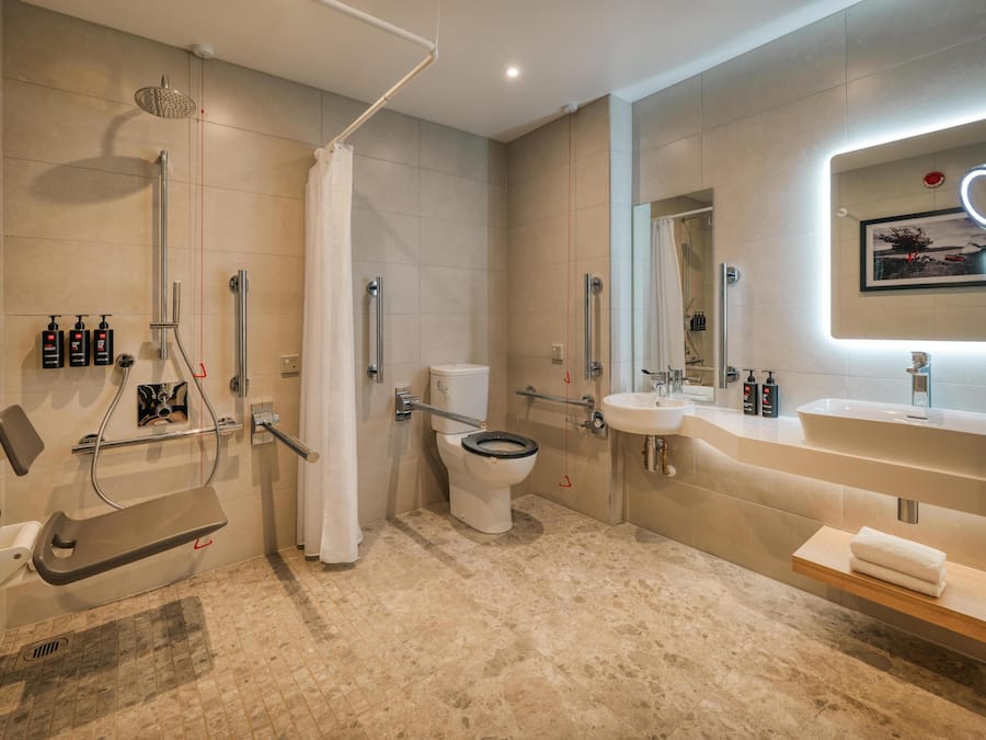 A modern bathroom with a shower, toilet, and sink, equipped with grab bars and a hand dryer.