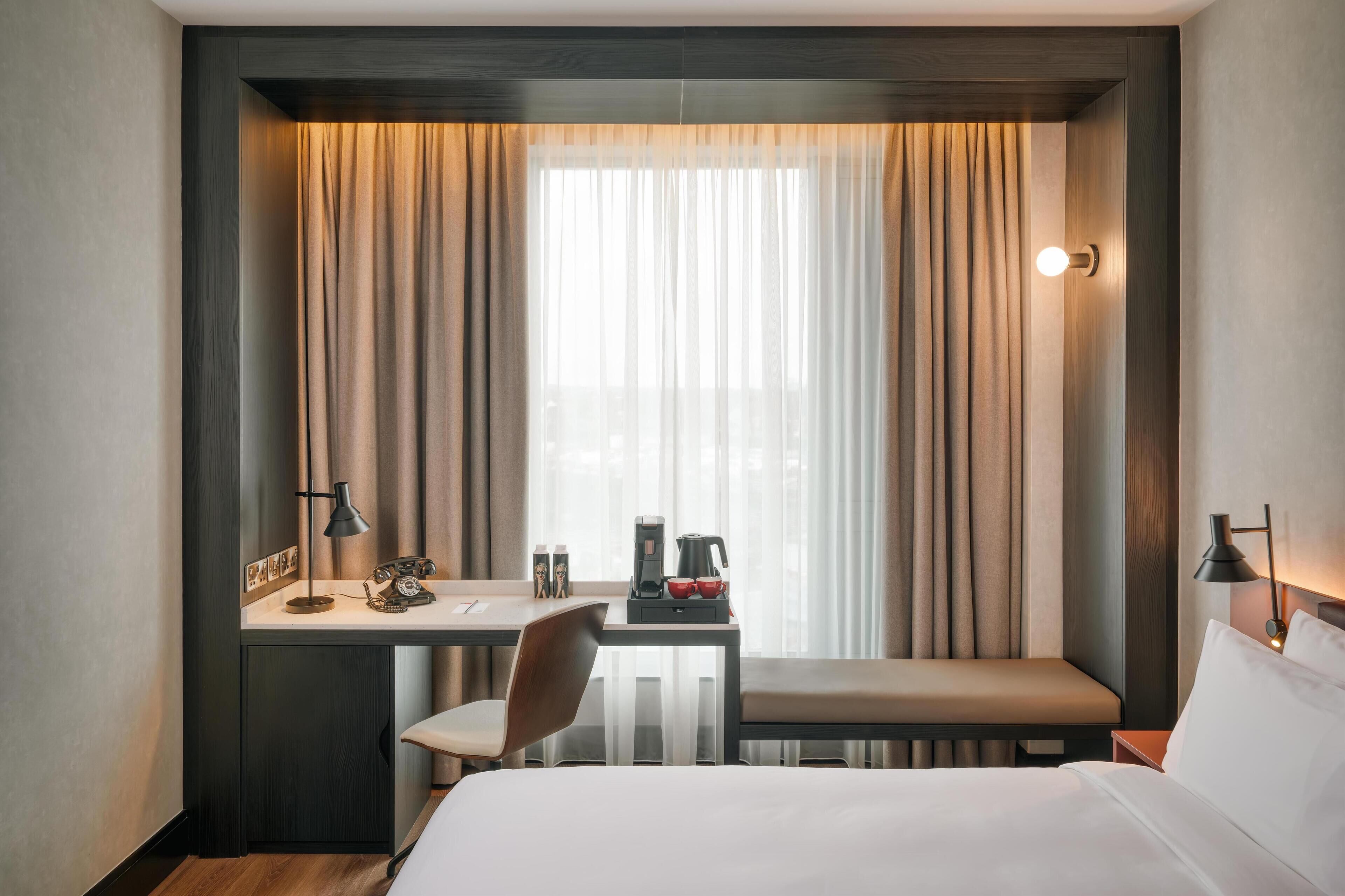 Una camera d'hotel con un letto, una scrivania con una sedia, un tavolino con una lampada, una panca e un'ampia finestra con tende.
