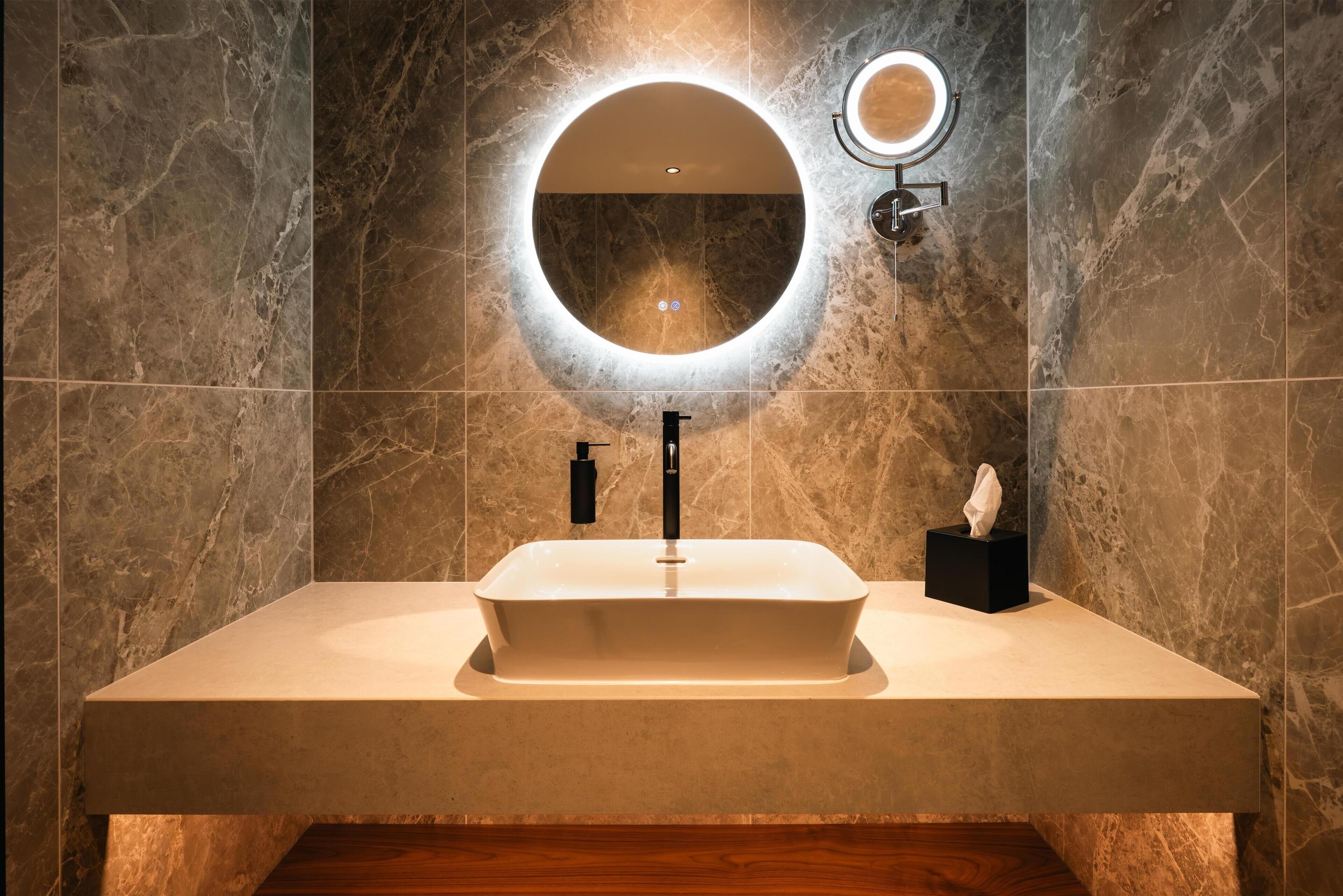 Un baño moderno con un lavamanos rectangular, un espejo redondo con luz y una caja de pañuelos en la encimera.
