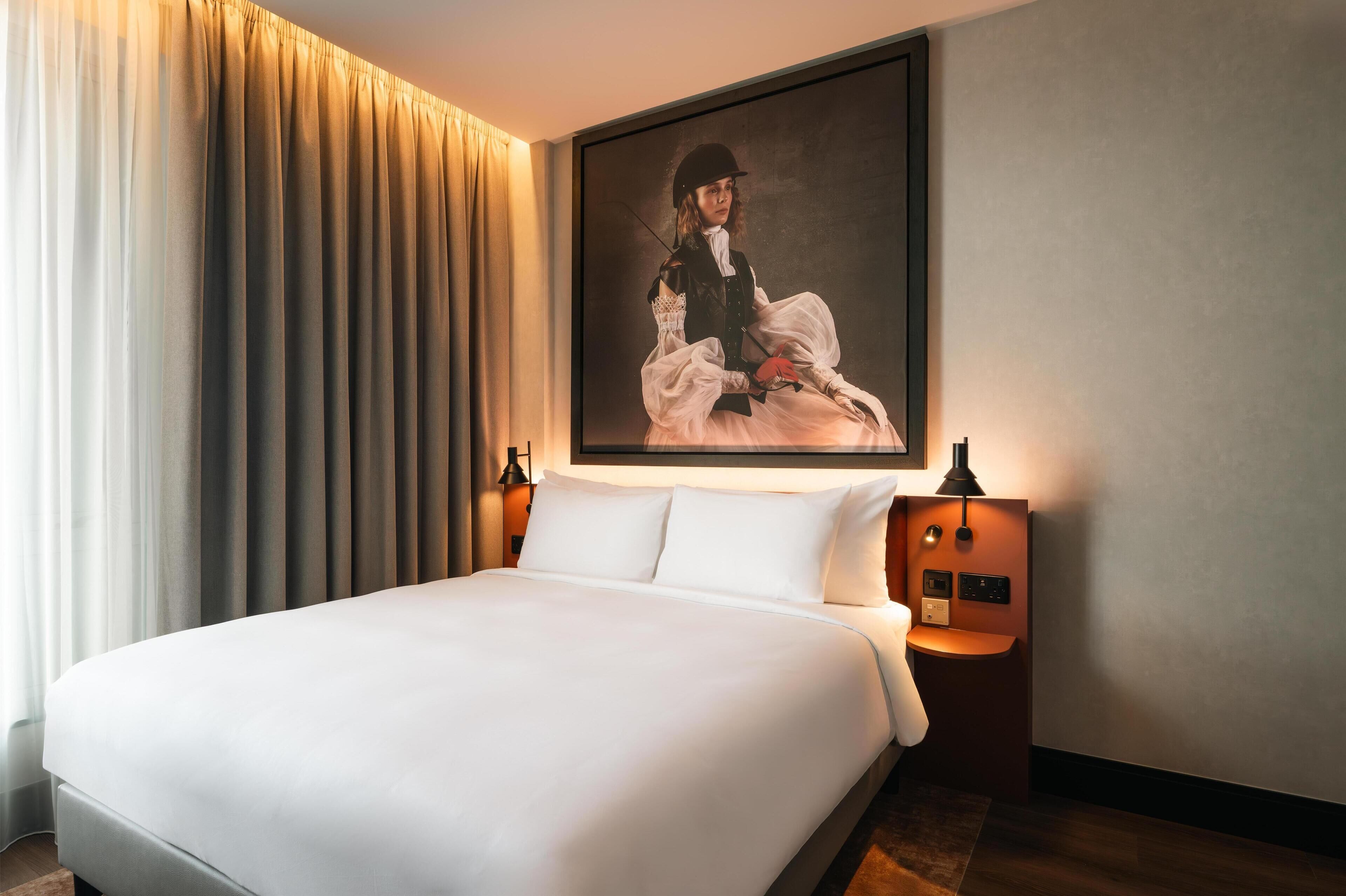 A hotel room with a large bed, a painting on the wall, and a nightstand with a lamp.