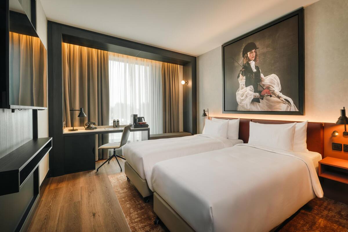 A hotel room with two beds, a desk, a chair, a desk lamp, a coffee maker, and a painting on the wall.