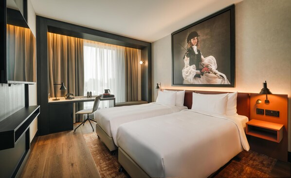 Habitación de hotel con dos camas, un escritorio con silla, un cuadro grande en la pared y una mesita de noche con lámpara.