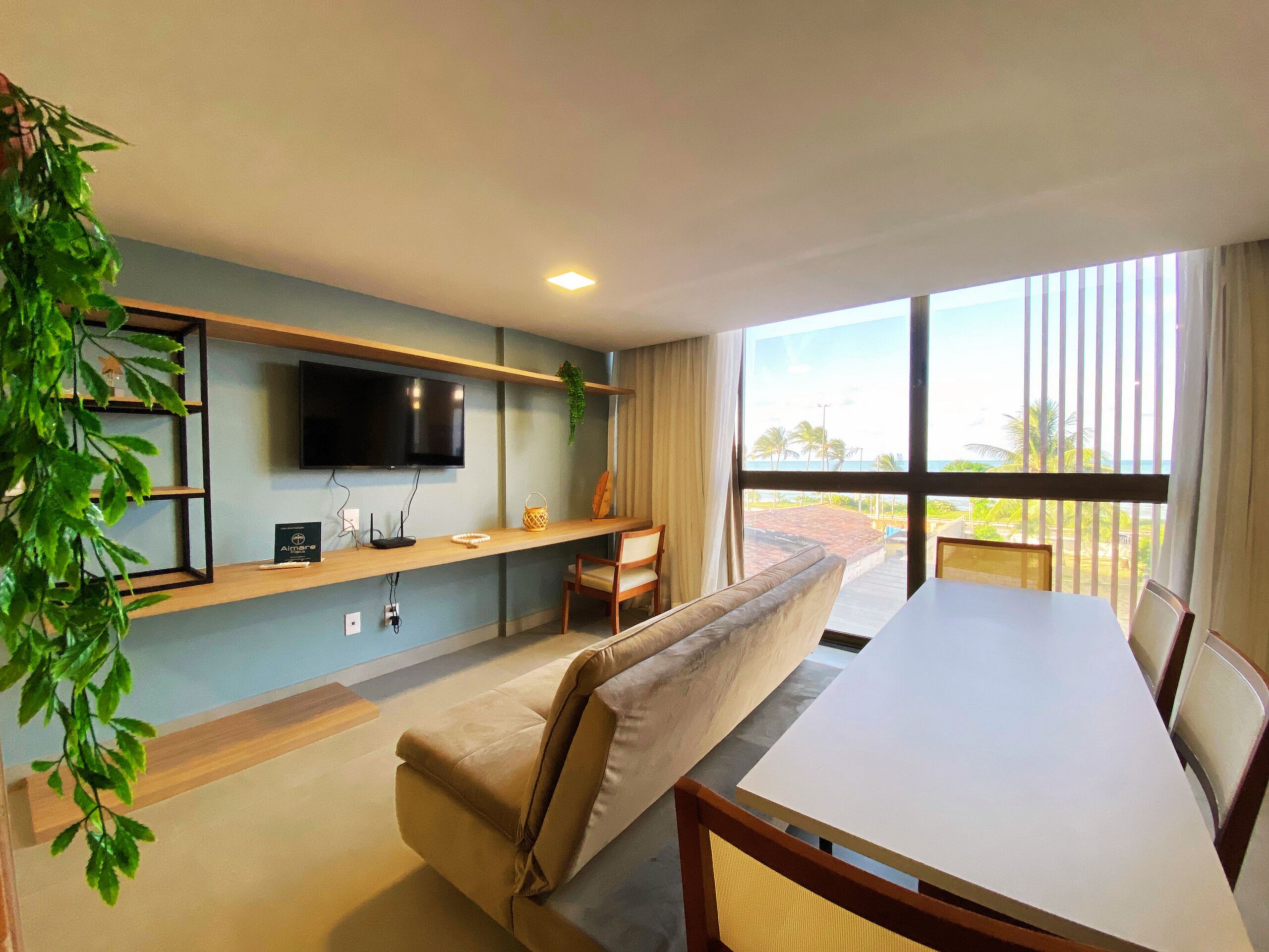 A modern living room with a flat-screen TV, a sofa, a dining table, and a large window offering a view of the outdoors.