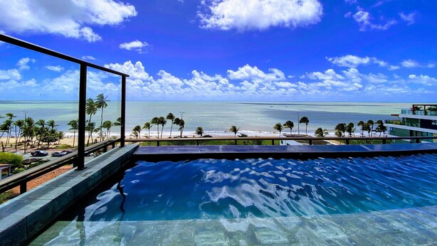 Una piscina con vistas al océano y a la playa, rodeada de palmeras y un cielo despejado.