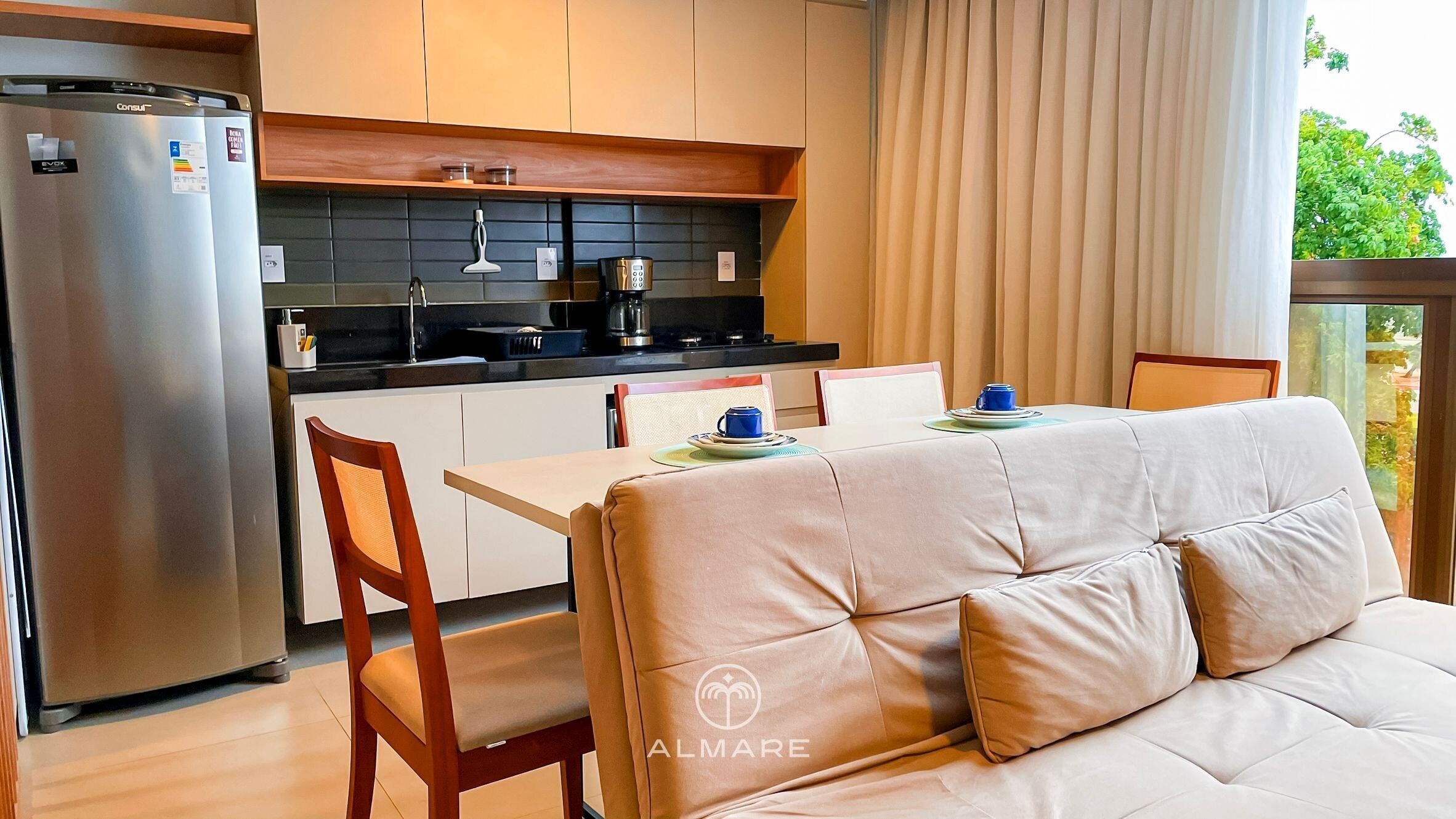A modern kitchen with a refrigerator, stove, and coffee maker. There is a dining table with chairs and a sofa.