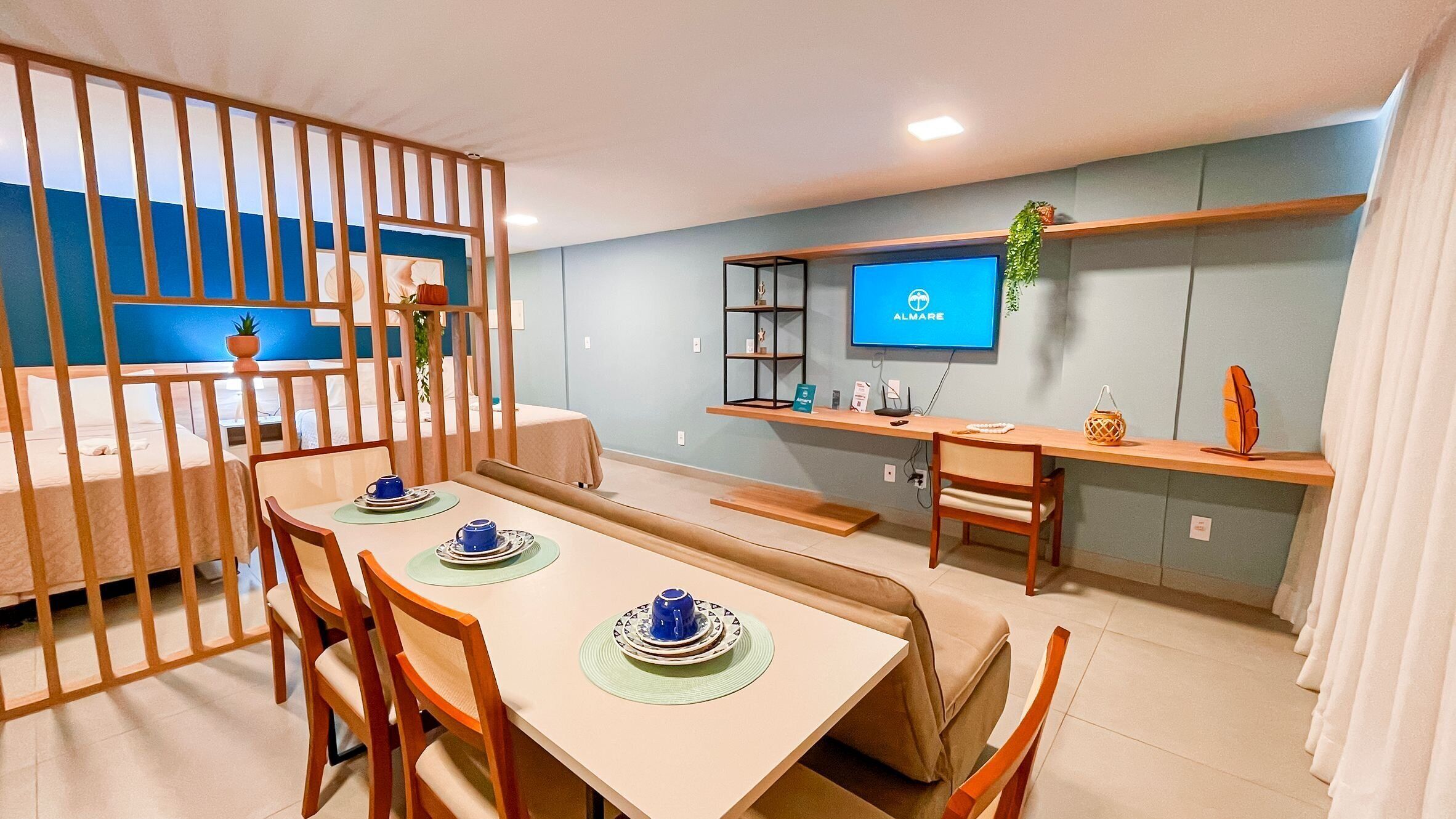 A modern hotel room with a dining area, a TV mounted on the wall, and a small shelf with decorative items.