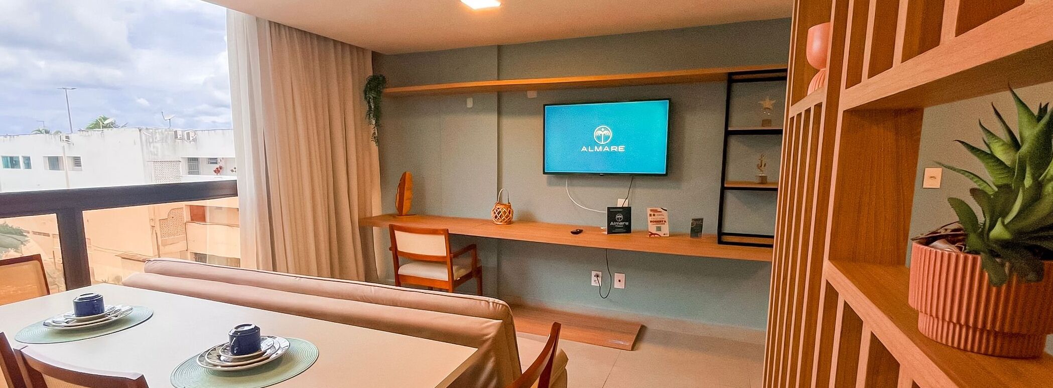 A modern living room with a flat-screen TV mounted on the wall, a wooden desk, and a dining area with a table set for two.