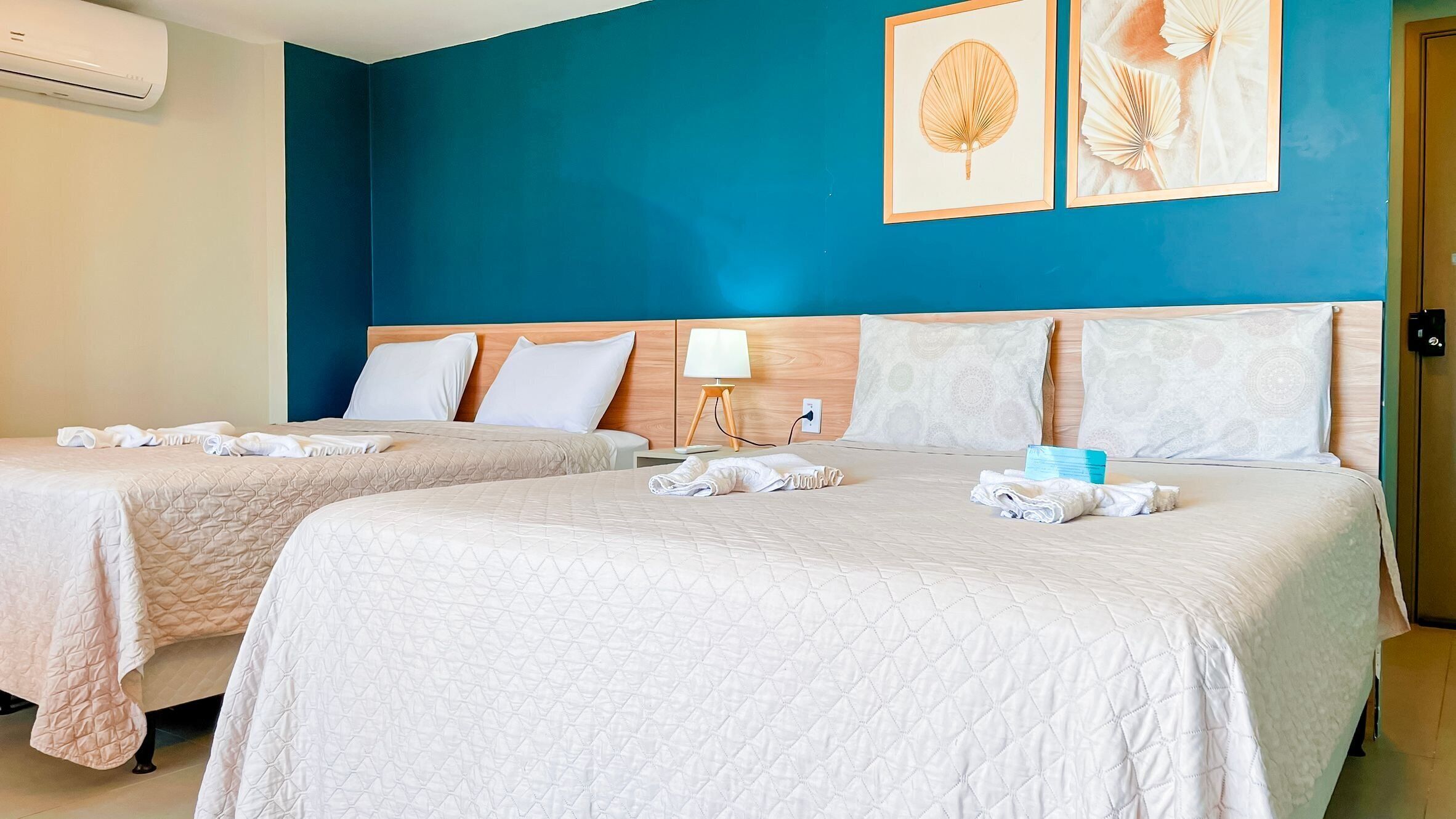 Habitación de hotel con dos camas, cada una con colcha acolchada, almohadas y toallas dobladas. Hay una mesita de noche con lámpara, un aire acondicionado de pared y dos cuadros enmarcados.