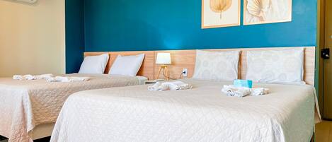 Habitación de hotel con dos camas, cada una con colcha acolchada, almohadas y toallas dobladas. Hay una mesita de noche con lámpara, un aire acondicionado de pared y dos cuadros enmarcados.