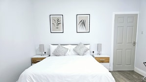 A modern bedroom with a large bed, two bedside tables, a nightstand, a wooden floor, and a door. - Mountpanther Holiday House (Downpatrick)