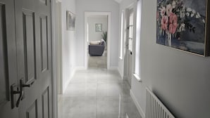 A hallway with white walls and doors, a tiled floor, a gray sofa, and a floral painting on the wall. - Mountpanther Holiday House (Downpatrick)