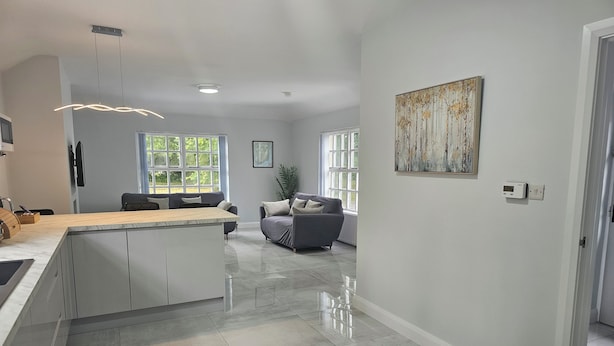 A modern kitchen with a central island, a dining area with a gray sofa, and a large window providing natural light. - Mountpanther Holiday House (Downpatrick)