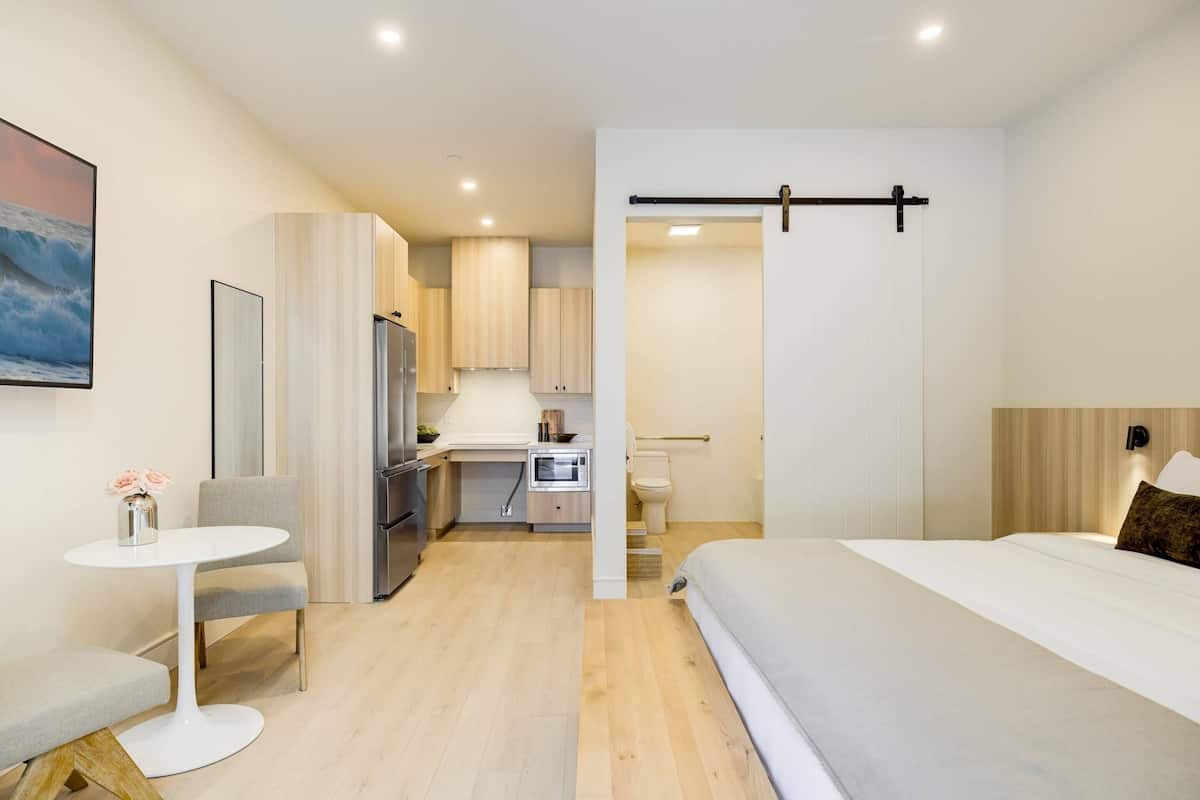 Ein modernes Hotelzimmer mit Bett, kleinem Schreibtisch, Stuhl und einer Küchenzeile.