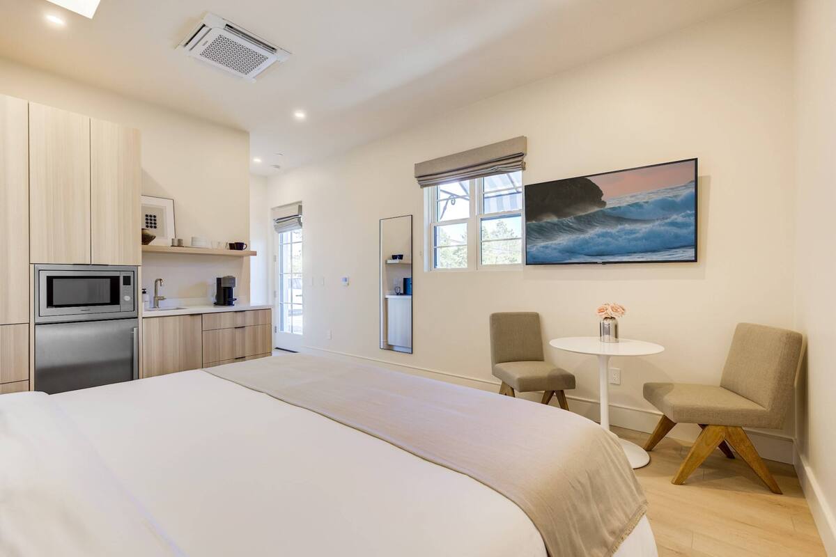 Ein modernes Hotelzimmer mit einem großen Bett, einer Küchenzeile, einer Essecke mit zwei Stühlen und einem Meeresblick-Gemälde.