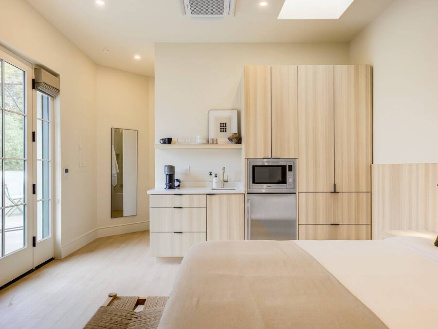 A modern bedroom with a large bed, a built-in wardrobe, a small kitchenette, and a sliding glass door leading to an outdoor area.