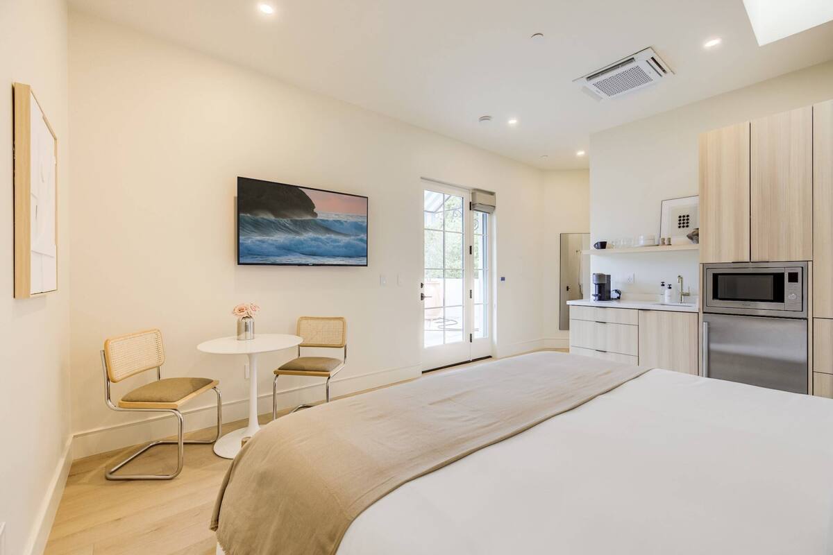Ein modernes Hotelzimmer mit einem großen Bett, einem kleinen Esstisch, zwei Stühlen, einer Küchenzeile mit Mikrowelle und Kühlschrank sowie einem Bild an der Wand.