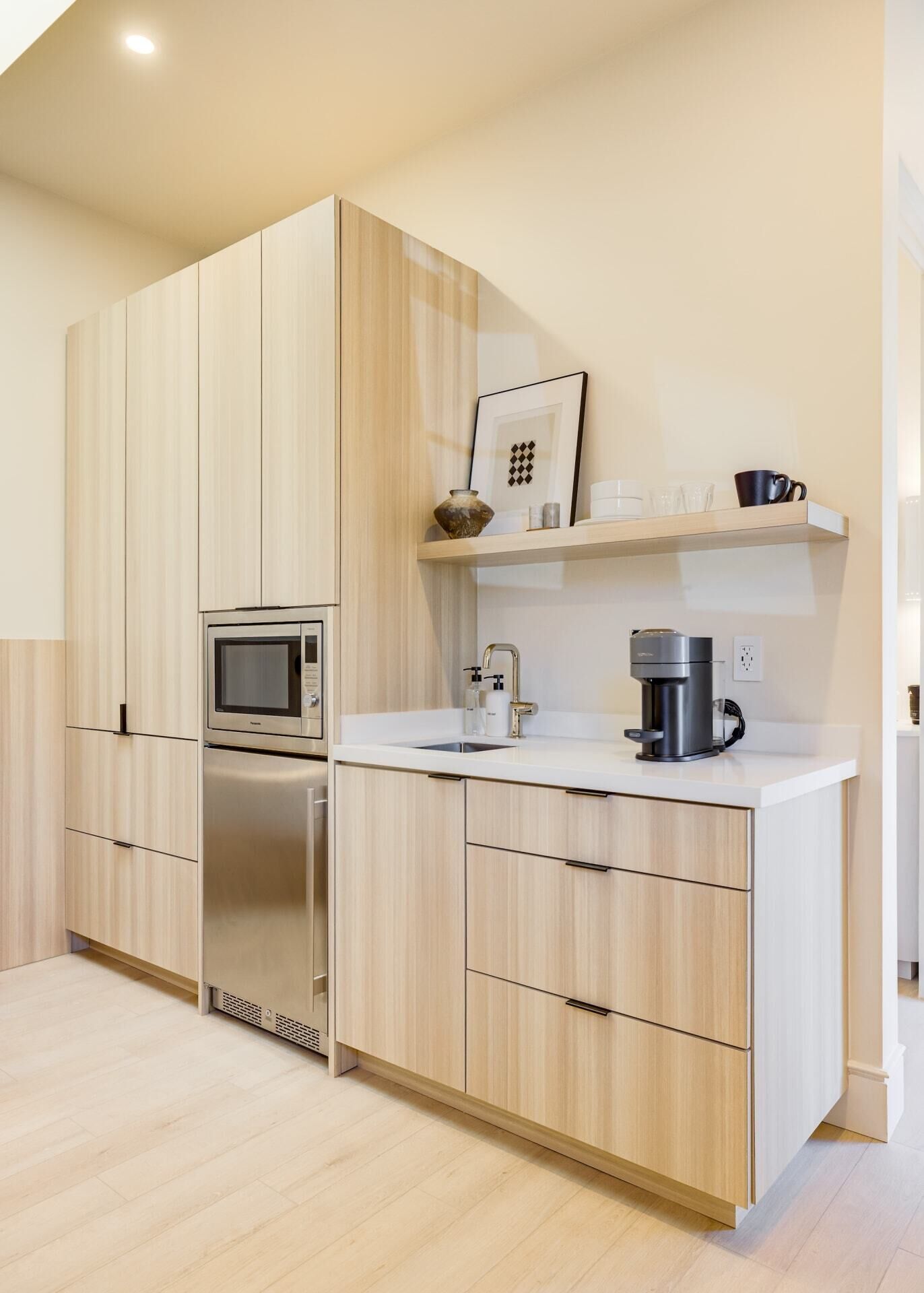 A modern kitchen with light wood cabinets, stainless steel appliances, a white countertop, and a small shelf with cups and a picture frame.