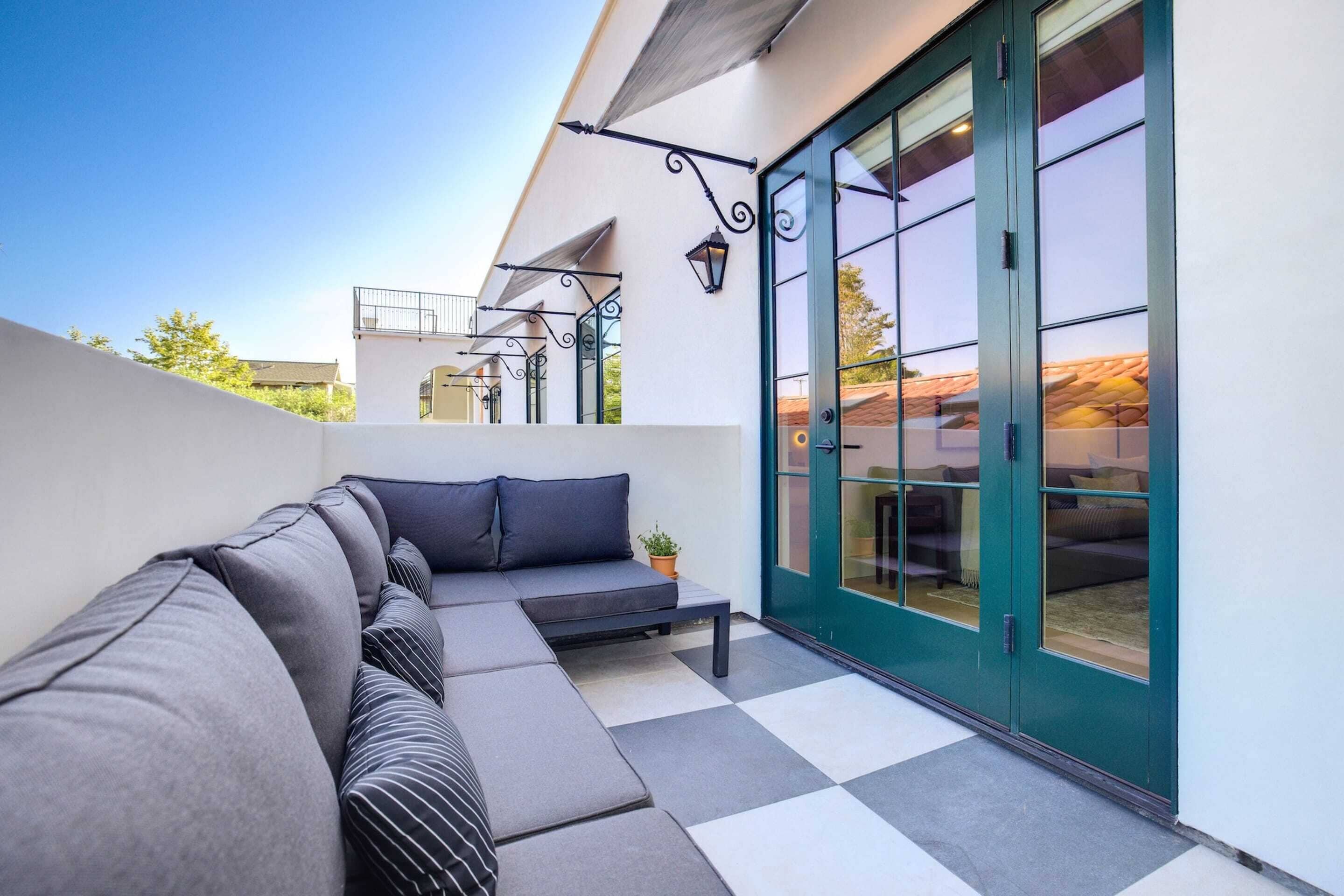 A modern outdoor seating area with a sofa and a bench, a glass door leading to an interior space, and a metal railing on a balcony.