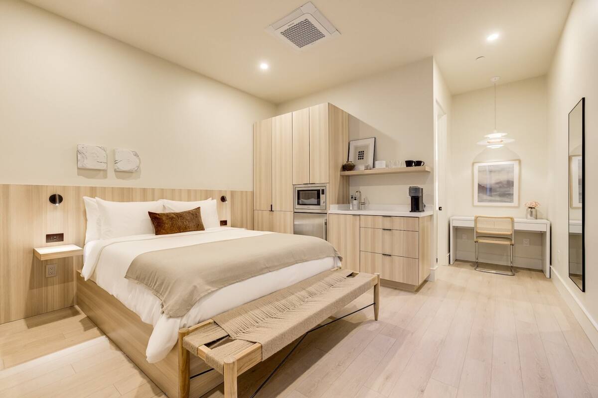 Ein modernes Schlafzimmer mit einem großen Bett, einem Holz-Kopfteil, Nachttischen, einer Bank, einem Einbauschrank, einer Küchenzeile und einem Schreibtisch mit Stuhl.