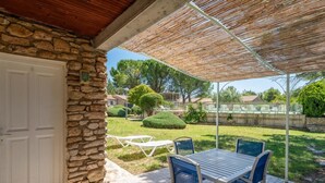 Un porche cubierto con paredes de piedra, una mesa y sillas, y vistas a un jardín cuidado y una cancha de tenis.