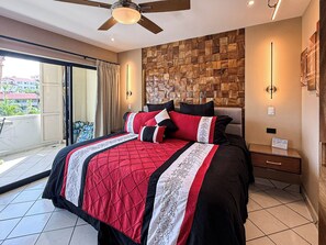 A hotel room with a bed, a ceiling fan, a wooden wall panel, a nightstand, a lamp, and a view of the outside. - A-303 Enjoy Your Vacation In This Delightful Remodeled Condominium! (Cabo San Lucas)