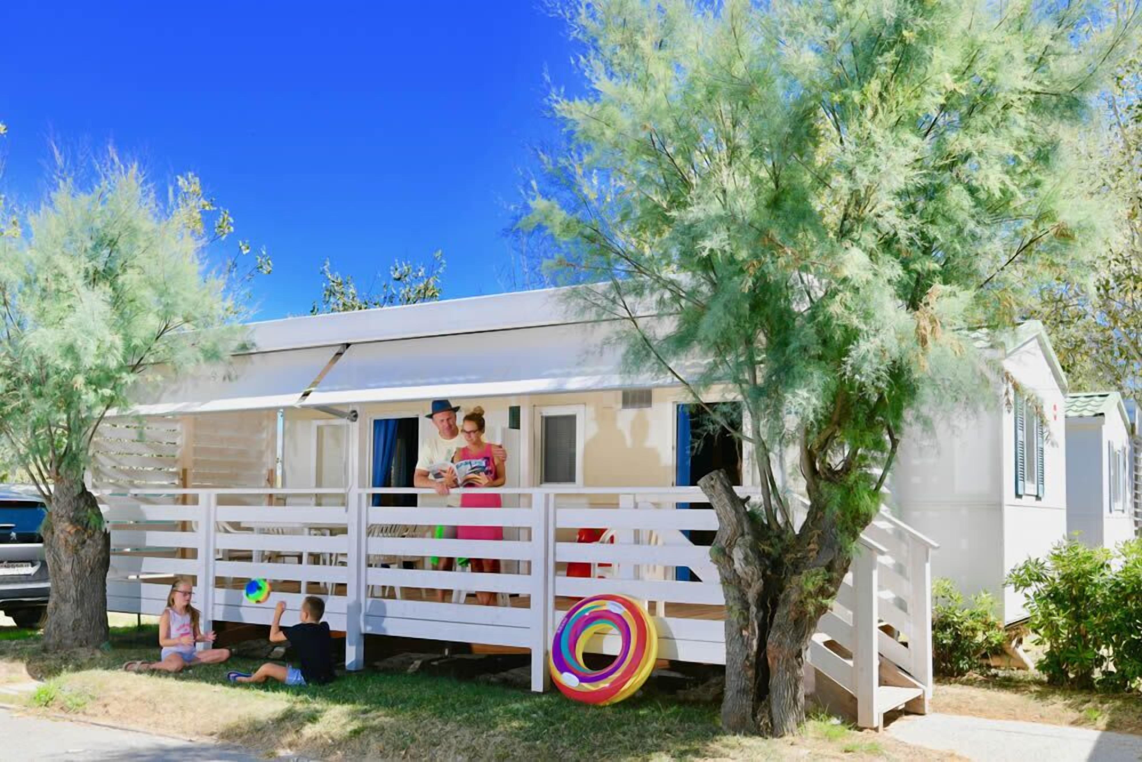 Une petite maison mobile blanche, dotée d’une véranda et entourée d’arbres.