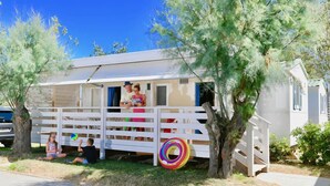 A small, white mobile home with a porch, surrounded by trees.