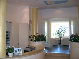 Un área de recepción con un mostrador curvo, plantas en macetas, una puerta blanca y vistas a una sala de estar con un ventanal grande.