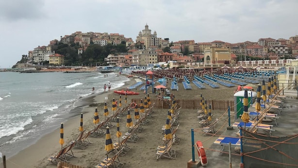 A beach with rows of sun loungers and umbrellas, a lifebuoy, and a view of a coastal town with buildings and a hill. - Robinia (Imperia)