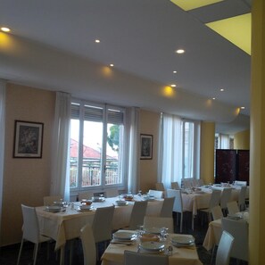 Une salle à manger avec des tables dressées pour le service, de grandes fenêtres avec des rideaux et des œuvres d’art encadrées accrochées aux murs.