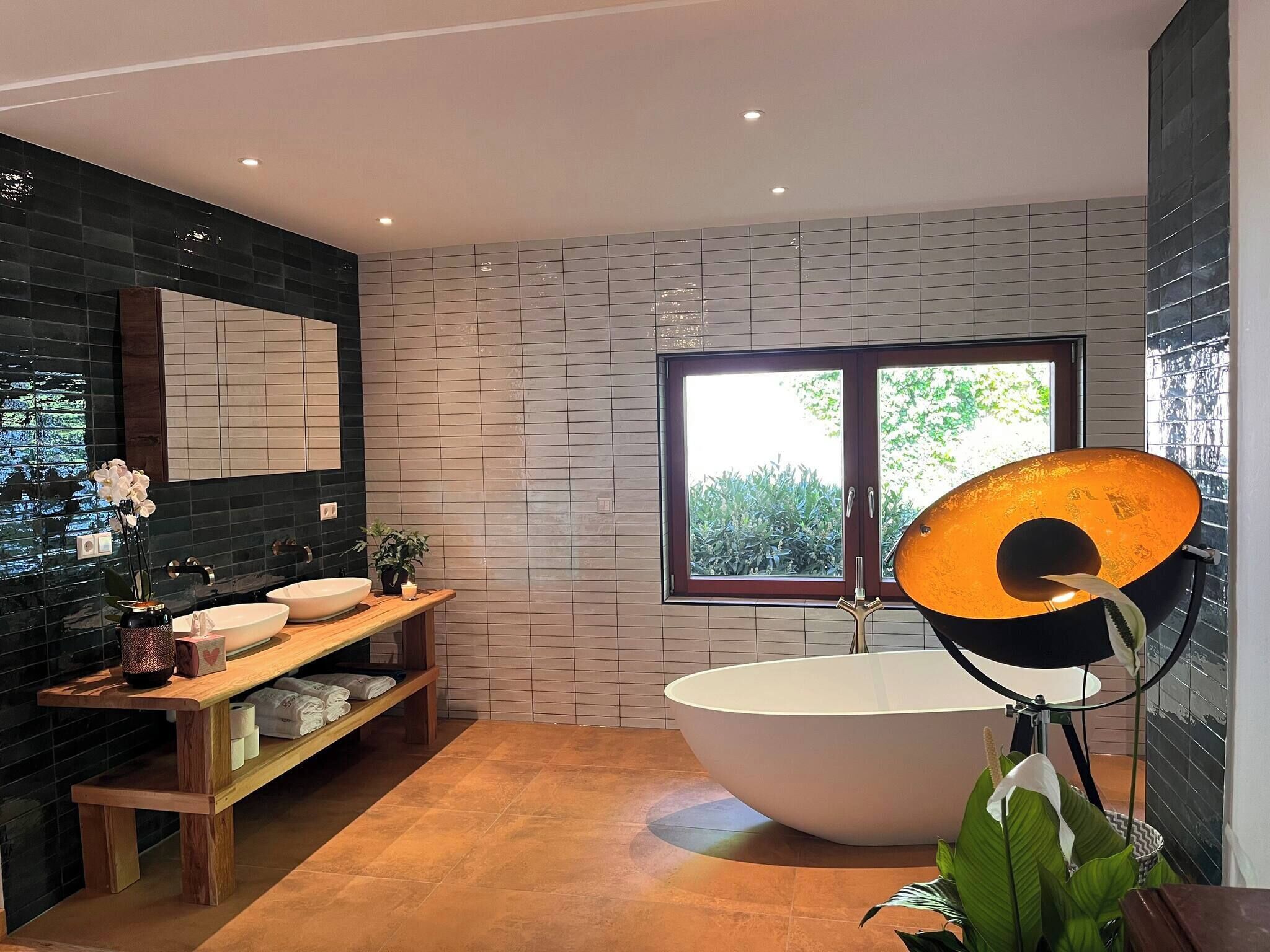 Une salle de bain moderne avec une baignoire sur pieds, un meuble-lavabo avec un miroir, une étagère avec des articles de toilette et une fenêtre donnant sur un espace verdoyant.