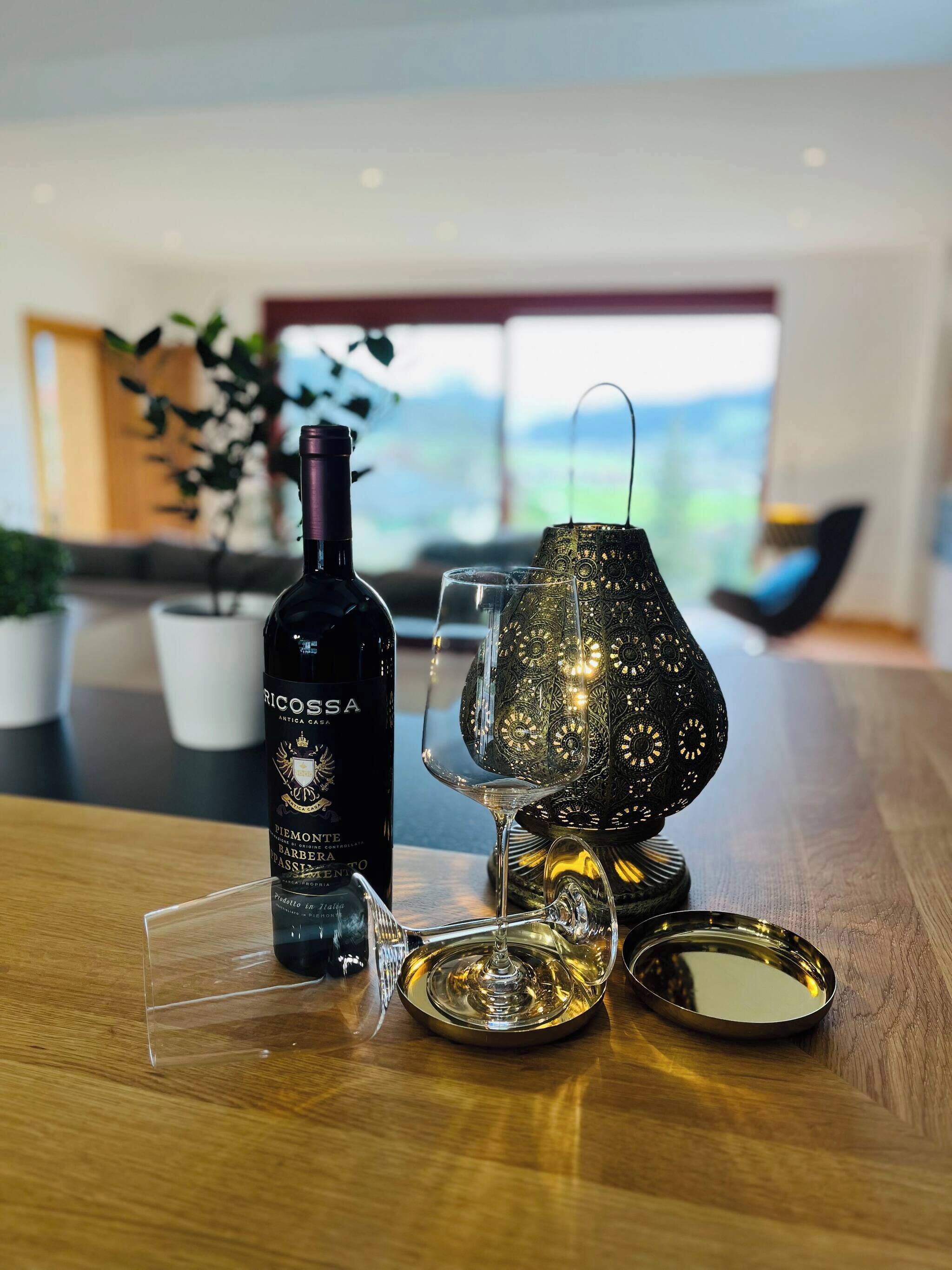 Una botella de vino, una copa de vino y una lámpara decorativa sobre una mesa de madera, con vistas a un paisaje a través de una ventana.