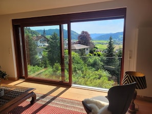 A room with a large sliding glass door leading to a scenic view of a village, a wooden table, a chair, and a lamp. - Graceland In the Black Forest (Fischerbach)