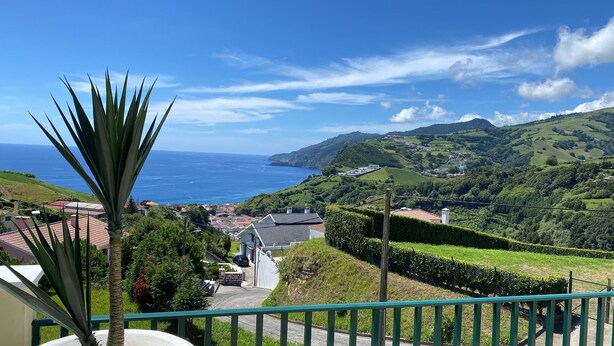 A coastal landscape with a view of the sea, rolling hills, and a residential area with houses and a paved road. - Vista do Topo- Apartment (Povoacao)