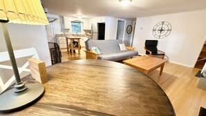 Un salon moderne avec une table en bois aux lignes courbes, un canapé et une table basse. On aperçoit à l’arrière-plan un coin cuisine avec une table à manger et des chaises.