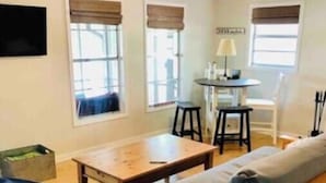 Un salon chaleureux avec un canapé en bois, une table basse ronde et un pouf bleu. Il y a deux fenêtres avec des stores marron, une petite table avec une lampe et un téléviseur fixé au mur.