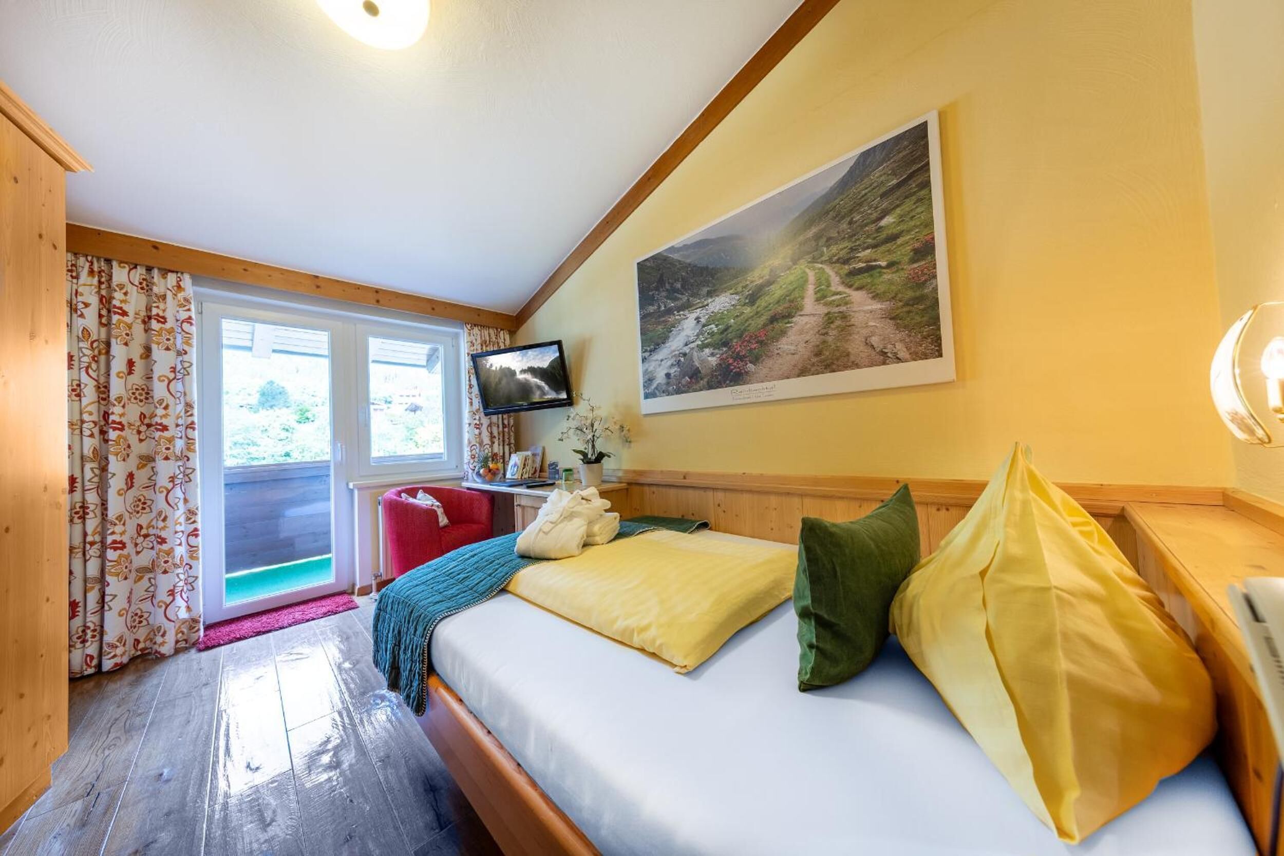 A cozy bedroom with a wooden bed, a television, a painting of a mountain landscape, and a window with curtains.