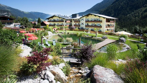 Hotel com várias varandas, um jardim com lago e montanhas ao fundo.