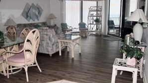 A spacious living room with a wooden floor, a glass coffee table, wicker chairs, and a view of the ocean through large windows.
