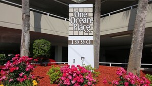 A building with a sign reading "One Ocean Place 1990" surrounded by red mulch and pink flowers.