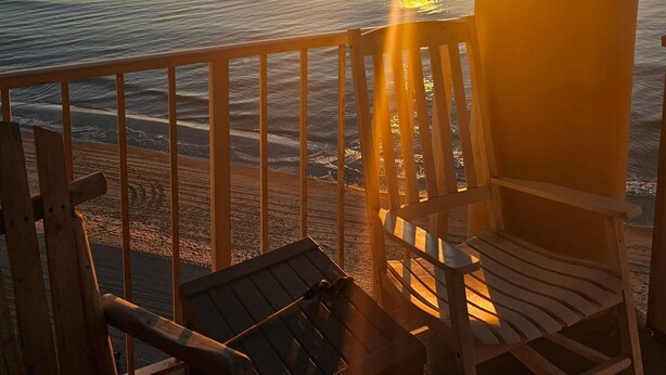 A wooden deck with two chairs and a table, overlooking a calm sea at sunset.