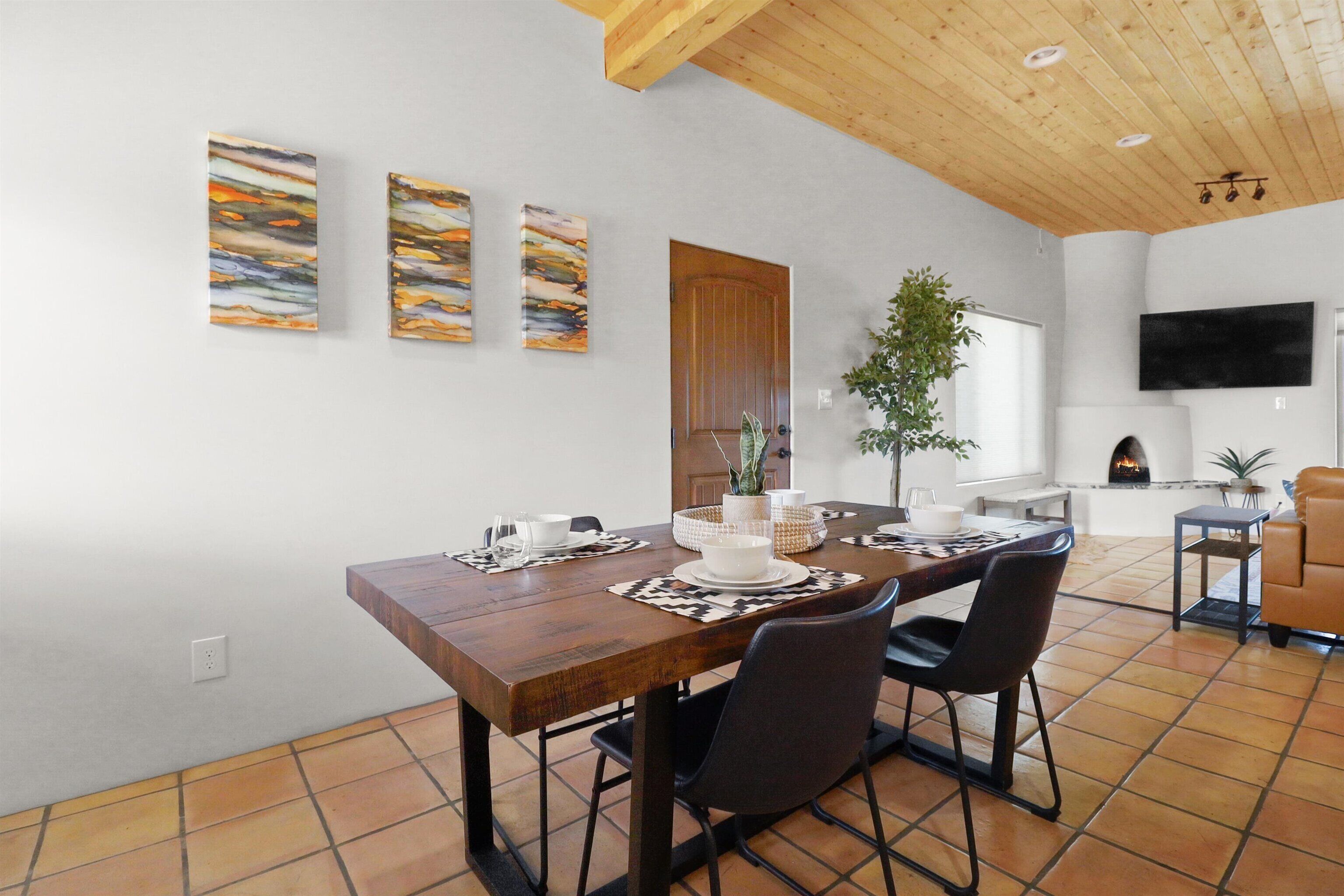 A dining area with a wooden table set for two, surrounded by chairs. There are three abstract paintings on the wall, a wooden door, and a television mounted on the wall.