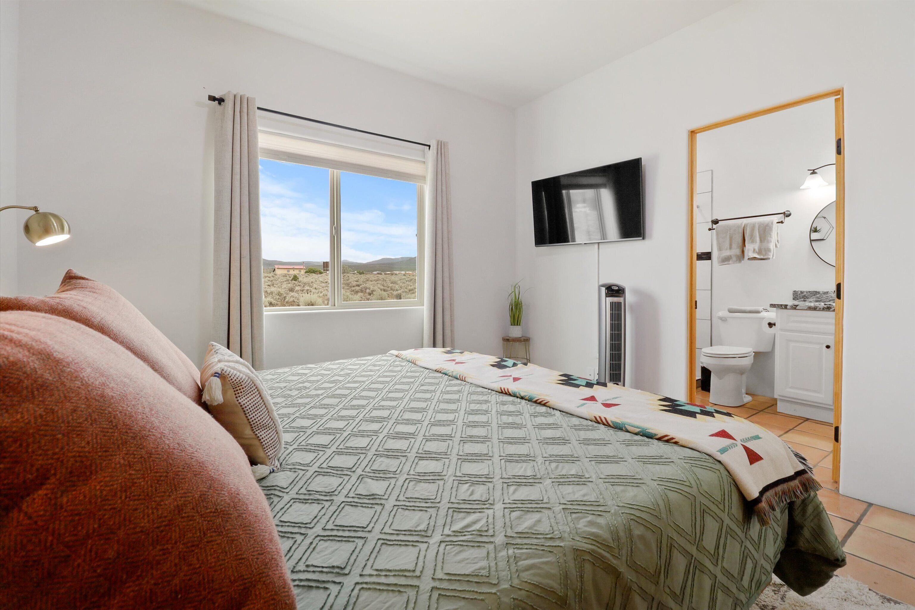 Ein Schlafzimmer mit Bett, einem an der Wand montierten Fernseher, einem Fenster mit Vorhängen und Blick auf die Wüste.