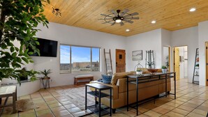 A spacious living area with a wooden ceiling, a large window, a sofa, a coffee table, and a TV mounted on the wall.