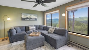 A modern living room with a sectional sofa, a ceiling fan, and a large window offering a view of the mountains. - New Hampshire vacation getaway with gorgeous mountain views—ski, hike, swim! (Thorton)