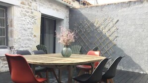 Une terrasse en bois avec une table ronde et des chaises, un vase rempli de fleurs et un mur de pierre avec une fenêtre.