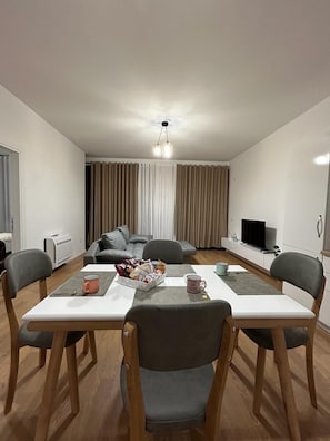 A modern living room with a dining table, two grey chairs, a sofa, a TV, and a ceiling light.
