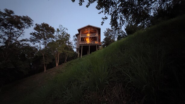 Une structure en forme de tour, faite de bois, avec un balcon, entourée d’arbres et d’herbe.